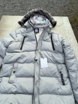 Ljusgrå dunjacka från Canada Goose - Snygg ljusgrå dunjacka från Canada Goose med avtagbar huva och fuskpäls. Jackan har två bröstfickor med dragkedja, två sidofickor med knappar och tydlig logga på ärmen. Perfekt för kalla vinterdagar och riktigt varm.