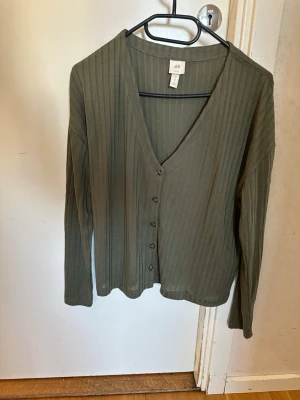 Olivgrön ribbad kofta från H&M - Olivgrön kofta med ribbad struktur och v-ringning. Långa ärmar och knäppning framtill med fem knappar. Tillverkad i mjukt material som känns skönt mot huden. Perfekt för lager på lager och enkel att styla.