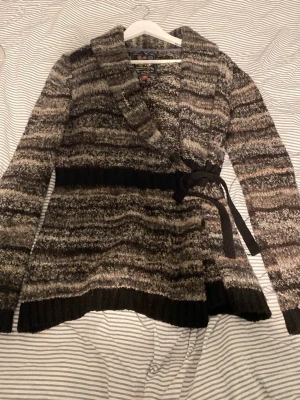 Randig kofta från Bondelid, M - Mysig stickad kofta från Bondelid i storlek M. Koftan har ett randigt mönster i bruna, grå och svarta toner, omlottmodell med knytband i midjan och bred krage. Perfekt för lager på lager och höstmys.