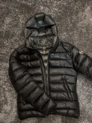 Colmar jacka - Snygg svart dunjacka från Colmar med huva och dragkedja framtill. Jackan har två sidofickor med dragkedja och Colmar-logga på vänster ärm. Perfekt för kalla vinterdagar tack vare det vadderade materialet. Se sista bilden för defekt!