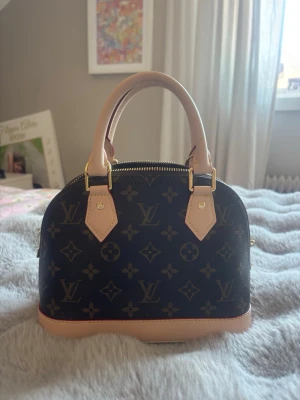 Alma BB Monogram handväska - Säljer en klassisk Alma BB Louis Vuitton handväska i monogram canvas med beige läderdetaljer och guldfärgade metalldelar. Köpt för drygt 1.5 år sen och har inte använts så mycket. Får med dustbag och box! 