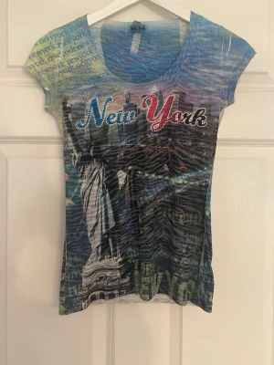  New York t-shirt med tryck - Cool t-shirt med New York-tryck och frihetsgudinnan framtill. Flera färger i blått, gult och rött, samt textdetaljer över hela tröjan. Kortärmad och figurnära modell, perfekt för dig som gillar unika motiv och streetstyle.