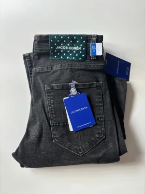 Svarta jeans från Jacob Cohën - Snygga svarta jeans från Jacob Cohën med raka ben och klassisk femficksdesign. Jeansen har en grön etikett med vita prickar bak och blåa tags framtill. Perfekta för dig som vill ha en stilren och modern look.