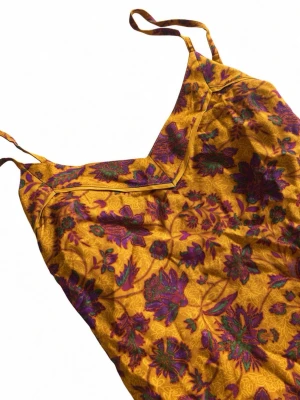 Gul blommig linne med smala band - Snygg gult linne. med lila och grönt blommönster. Klänningen har smala axelband och v-ringad halsringning. Perfekt för varma dagar och ger en färgstark och bohemisk vibe. Materialet känns lätt och luftigt.