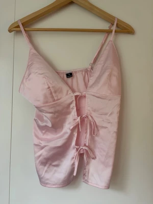 Ljusrosa satinlinnetopp med snörning - Säljer en ljusrosa linnetopp i satin med tunna axelband och snygg snörning framtill. Toppen har en djup v-ringning och croppad passform som ger en trendig look. Perfekt att styla med jeans eller kjol för en cool outfit.