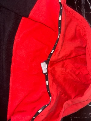 Svart och röd Puma hoodie - Säljer en snygg svart hoodie från Puma med röda detaljer och huva. Hoodien har dragkedja framtill, fickor på sidorna och Puma-loggor tryckta över bröstet. Perfekt för dig som gillar sportig stil och vill sticka ut lite extra.