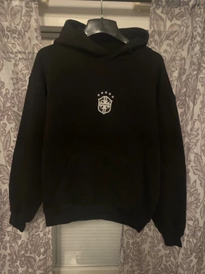 Svart hoodie med vit logga - Svart hoodie med huva och en vit logga på bröstet. Klassisk känguruficka framtill och ribbade muddar vid ärmslut och nederkant. Perfekt för en chill och avslappnad stil.