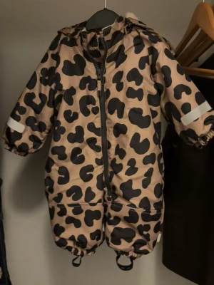 Leopardmönstrad overall med huva - Säljer en beige overall med svart leopardmönster och huva. Overallen har dragkedja framtill, resår vid ärm- och benslut samt reflexdetaljer på armarna. Perfekt för kyligare dagar och enkel att ta på tack vare dragkedjan.