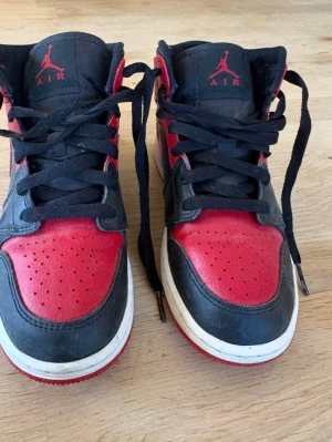 Svarta och röda Air Jordan 1 Mid - Säljer ett par Air Jordan 1 Mid sneakers i svart och rött läder med klassisk Nike Swoosh på sidan och Air Jordan-logga. Skorna har svart snörning, perforerad tåbox och vit mellansula med röd yttersula. Perfekta för dig som gillar streetwear och ikonisk design.