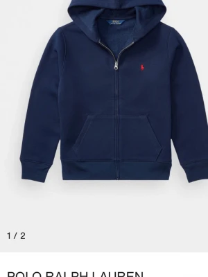Marinblå hoodie från Polo Ralph Lauren - Snygg marinblå hoodie med dragkedja från Polo Ralph Lauren. Klassisk röd logga broderad på bröstet, vita dragsnören i huvan och känguruficka framtill. Tillverkad i mjukt bomullsmaterial som är skönt att ha på sig. Perfekt för en avslappnad och stilren look.