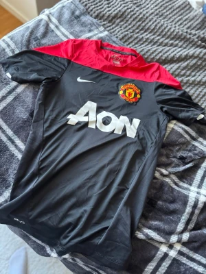 Manchester United Nike matchtröja - Snygg Manchester United matchtröja från Nike i svart med röd axelparti och vit logga. Tröjan har klubbmärke på bröstet, AON som sponsortryck och är tillverkad i lätt, andningsbart material med Dri-FIT-teknologi. Perfekt för fotbollsträning eller att bära till vardags.