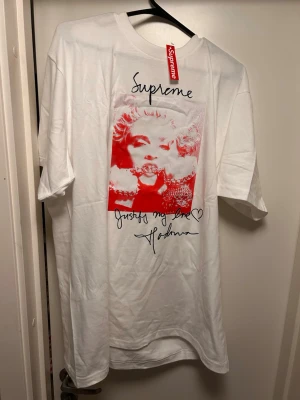 Supreme x Madonna vit t-shirt - Supreme t-shirt i vitt med stort rött och vitt tryck av Madonna på framsidan. Trycket har Supreme-loggan upptill och Madonnas autograf samt texten 'Justify my love' nedtill. Klassisk passform och rund hals. Perfekt statement-plagg för streetwear.