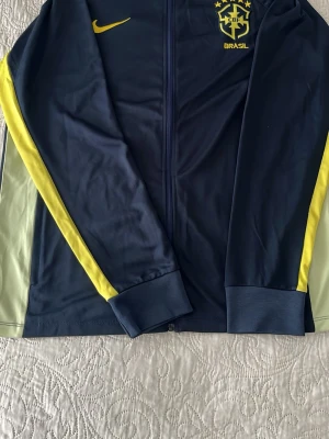 Blå Brasil träningsjacka Nike - Snygg blå träningsjacka från Nike med Brasil-logga och gula detaljer längs ärmarna. Jackan har hel dragkedja framtill och ribbade muddar. Perfekt för fotbollsträning eller som sportig vardagsstil.