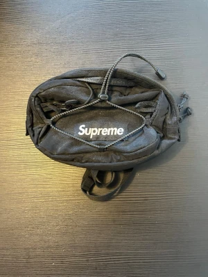 Svart magväska från Supreme - Supreme magväska i svart nylon med justerbara remmar och dragkedja. Väskan har ett stort huvudfack, mindre ytterfack och elastisk snörning på framsidan. Perfekt för dig som vill ha en streetwear-look och praktisk förvaring.