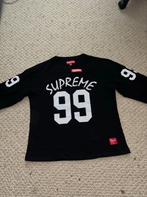 Svart Supreme topp med 99-tryck - Supreme långärmad svart topp med stora vita siffror '99' och Supreme-tryck både fram och bak. Tröjan har sportig vibe och är tillverkad i mjuk bomull. Perfekt för dig som gillar streetwear och vill sticka ut.