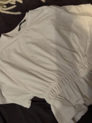 Vit topp med resår i midjan - Säljer en vit topp med korta ärmar och rund halsringning. Toppen har en snygg resårdetalj i midjan som ger en fin form och lite volang nedtill. Perfekt att styla med jeans eller kjol för en fräsch look.