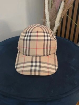 Burberry keps - Snygg beige keps från Burberry med klassiskt rutmönster i svart, vitt och rött. Justerbar baktill med svart spänne och tydlig Burberry London England-logga. Perfekt accessoar för en trendig och stilren look.