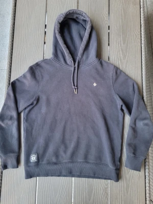 Morris hoodie - Säljer en mörkblå hoodie med dragsko. Tröjan har en liten broderad logga på bröstet och en patch nertill vid midjan. Klassisk passform och ribbade muddar vid ärmslut och nederkant. Perfekt för en avslappnad stil.