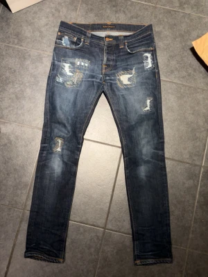 Blå nudie jeans med patchdetaljer - Säljer ett par riktigt snygga blå jeans med slitningar och patchdetaljer på låren och knäna. Jeansen har smal passform och klassisk femficksdesign. Perfekta för dig som gillar en trendig och avslappnad stil.