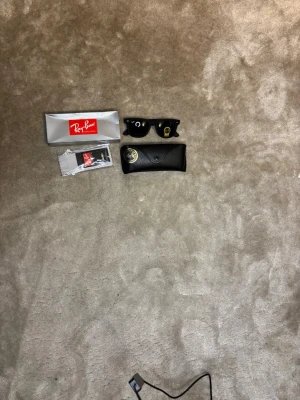 Svarta Ray-Ban Wayfarer solglasögon - Klassiska svarta Ray-Ban Wayfarer solglasögon med ikonisk form. Kommer med originalfodral, putsduk och förpackning. Snygg och tidlös design som passar till allt. Perfekta för soliga dagar och ger en cool vibe. Fick i present vet ej om real.