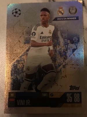 Match Attax Real Madrid kort Vini Jr. - Samlarkort från Topps Match Attax med Vini Jr. i Real Madrid C.F. Kortet har guldiga och glittriga detaljer, Champions League-märke och Real Madrids logga. Perfekt för dig som gillar fotboll och samlarkort.