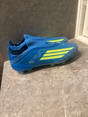 Blåa Adidas F50 fotbollsskor - Säljer ett par blåa Adidas F50 fotbollsskor med neon-gula detaljer och klassiska tre ränder på sidan. Skorna har en strömlinjeformad design utan snörning och är tillverkade i syntetmaterial för bästa grepp och känsla på planen.
