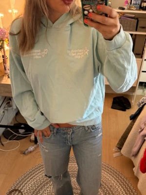 Ljusblå hoodie med tryck - Säljer en ljusblå hoodie med vit text framtill och stort tryck på ryggen. Hoodien har huva och ribbade muddar vid ärmar och midja. Perfekt för en avslappnad streetstyle-look. Materialet känns mjukt och bekvämt.