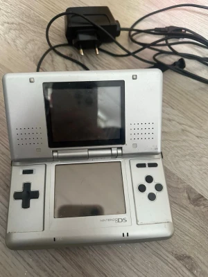Nintendo DS silver med laddare - Nintendo DS i silverfärg med dubbla skärmar, styrkors, knappar och touchskärm. Konsolen är hopfällbar och kommer med original laddare och handledsrem. Perfekt för portabelt spelande och klassiska DS-spel. 