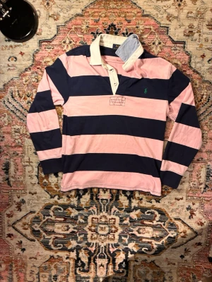 Randig rugbytröja i rosa och marinblå - Säljer en långärmad rugbytröja med breda rosa och marinblå ränder. Tröjan har klassisk krage i vitt och ljusblått samt knappar i halsen. Broderad grön logga på bröstet. Perfekt för en sportig och avslappnad stil.