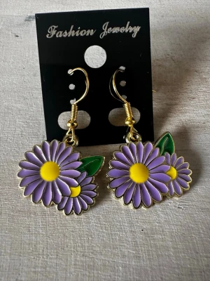 Lila blomörhängen med gulddetaljer - Snygga örhängen i form av lila blommor med gula mitten och gröna blad. Hängande modell med guldfärgade krokar och detaljer. Perfekta för dig som vill ha en färgglad och lekfull accessoar till din outfit.