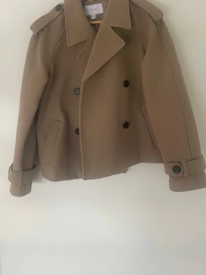 Kort beige kappa med dubbelknäppning - Stilren kort beige rock i ullmix med dubbelknäppning och bred krage. Rocken har axelklaffar, två snedställda fickor och dekorativa knappar vid ärmsluten. Perfekt för kyligare dagar och ger en klassisk look. Väldigt höstig och mysig. Kan verkligen fixa en outfit. Köpt här på Vinted men aldrig andvänd av mig. Lite nopprig men går säkert att fixa med en noppkamm. Annars i fint skick