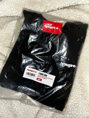 Svart t-shirt från Tyngre - Säljer en svart t-shirt från Tyngre med vit logotyp på bröstet. Klassisk rund hals och rak passform. Perfekt för dig som gillar enkel och stilren design. T-shirten är i dammodell och tillverkad i mjukt material.