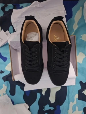 Christian Louboutin  - Snygga svarta sneakers från Christian Louboutin med klassisk röd sula och svart mockaliknande material. Skorna har svarta skosnören, låg profil och diskret logga på innersulan. Perfekta för dig som vill sticka ut med stilrena detaljer.