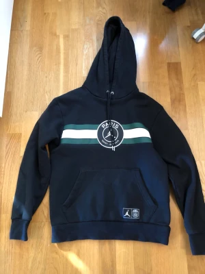 Jordan PSG svart hoodie med huva - Fin hoodie i bra skick förutom en mindre fläck på baksidan av ärmen. Har ej testat fläckborttagning så kan försvinna. Se bild. Bra kvalitet i tjockt tyg.