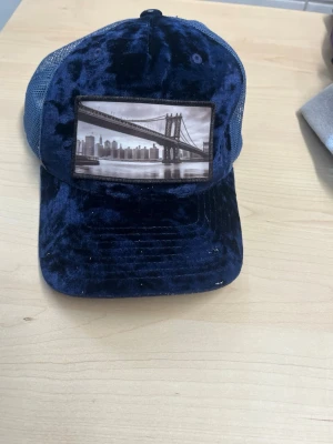 Blå sammetskeps med broderad bro - Snygg blå keps i sammetsmaterial med mesh på sidorna och bak. Framtill finns en broderad patch med motiv av en bro och stadssilhuett. Kepsen har böjd skärm och justerbar baksida för perfekt passform. Kepsen har en liten vit fläck.