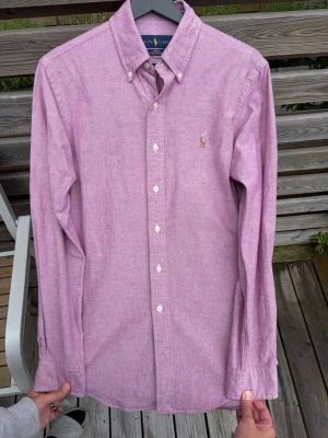 Ljuslila skjorta från Ralph Lauren - Säljer en ljuslila skjorta från Ralph Lauren med klassisk button-down krage och broderad logga på bröstet. Skjortan har vita knappar, lång ärm och är i slim fit. Perfekt för dig som gillar stilrena plagg med snygga detaljer.