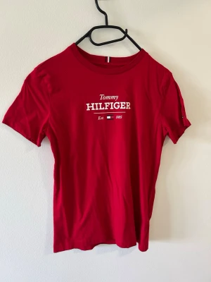Röd Tommy Hilfiger t-shirt - Snygg röd t-shirt från Tommy Hilfiger med klassisk logga och texten 'Est. 1985' framtill. T-shirten har rund halsringning och korta ärmar. Tillverkad i mjuk bomull som känns skön mot huden. Perfekt för en avslappnad och stilren look.