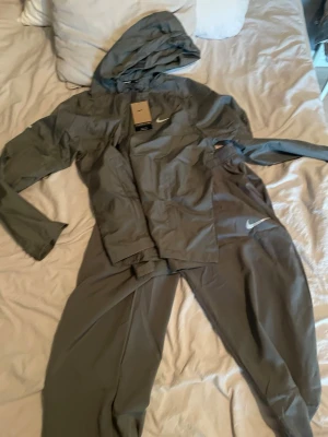 Grått Nike träningsset med huva - Säljer ett grått träningsset från Nike med matchande jacka och byxor. Jackan har huva, dragkedja och Nike-logga på bröstet. Byxorna har resår i midjan och Nike-logga på benet. Perfekt för löpning eller gym. Materialet är lätt och andas.
