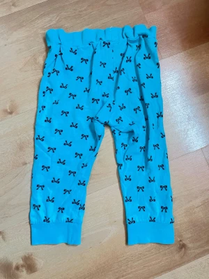 Blå leggings med svarta rosetter, 86/92 - Supersöta blå leggings med svarta rosett-mönster över hela tyget. De har en bekväm resår i midjan och muddar vid bensluten. Perfekta för en lekfull och färgglad stil. Materialet känns mjukt och stretchigt.