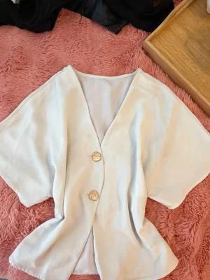 Vit kortärmad blus med knappar - Stilren vit blus med v-ringning och två dekorativa knappar framtill. Blusen har en lös passform och korta ärmar, vilket ger en avslappnad och trendig look. Tillverkad i ett mjukt, tunt tyg som känns svalt och bekvämt mot huden.