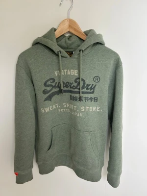 Grön Superdry hoodie med tryck - Snygg grön hoodie från Superdry med vintage-tryck på bröstet och japanska tecken. Klassisk känguruficka framtill, dragsko i huvan och ribbade muddar. Perfekt för en avslappnad streetstyle-look.