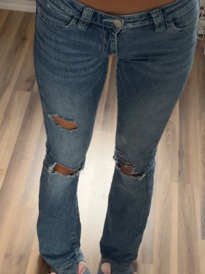 Blå slitna bootcut jeans - Säljer ett par blå bootcut jeans med slitningar och hål vid knäna. Jeansen har låg midja, klassiska fem fickor och dragkedja med knapp. Perfekta för en avslappnad och trendig look.