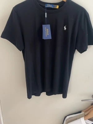 Svart t-shirt från Polo Ralph Lauren - Klassisk svart t-shirt från Polo Ralph Lauren med den ikoniska vita logon broderad på bröstet. T-shirten har rund halsringning och korta ärmar. Tillverkad i mjuk bomull för en skön känsla och enkel stil.