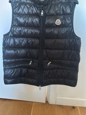 Svart dunväst från Moncler - Snygg svart dunväst från Moncler med glansig finish och quiltad design. Västen har två dragkedjeförsedda fickor framtill, hög krage och Moncler-logga på bröstet. Perfekt för lager-på-lager och håller dig varm med stil.