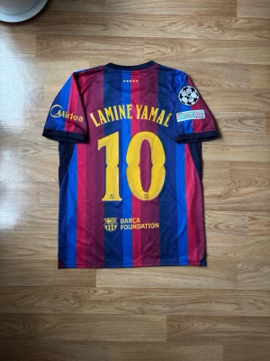 Barcelona matchtröja Lamine Yamal #10 | 2026/27 - Säljer en FC Barcelona matchtröja med Lamine Yamal och nummer 10 på ryggen. Tröjan har klassiska blå och vinröda ränder, gula detaljer och flera officiella loggor som Champions League och Spotify. Tillverkad i lätt och ventilerande material, perfekt för fotboll.
