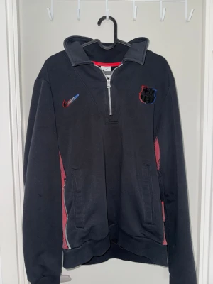 FC Barcelona Tröja med halvdragkedja  - Säljer en svart långärmad tröja från Nike med FC Barcelona-logga på bröstet och rödblå Nike swoosh. Tröjan har halv dragkedja, hög krage och röda mesh-detaljer på sidorna. Perfekt för träning eller chill. Två fickor med dragkedja framtill.