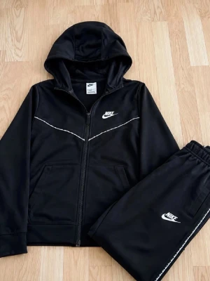 Nike Youth Black Tracksuit Set - This image shows a black Nike tracksuit consisting of a hooded zip-up jacket and matching pants. Both pieces feature the Nike logo in white. The tracksuit is laid out on a wooden floor.