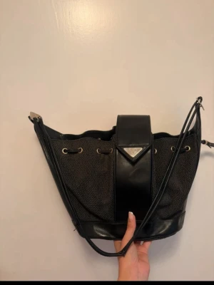 Vintage Yves Saint Laurent (YSL) Triangle Logo Bucket Bag - Snygg svart bucket bag med detaljer i läder och metall. Väskan har dragsko upptill, axelrem och en bred läderremsa med metallogo framtill. Perfekt för dig som gillar stilrena accessoarer med en twist.