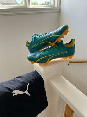 Gröna Puma fotbollsskor med gult - Snygga gröna fotbollsskor från Puma med gula detaljer och mönstrad ovandel. Skorna har gula dobbar och snörning för bra passform. Perfekta för dig som vill sticka ut på planen och ha bra grepp på gräs.