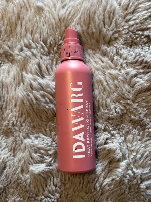 IDAWARG värmeskyddsspray rosa - Säljer en rosa värmeskyddsspray från IDAWARG. Flaskan är cylinderformad med spraymunstycke och vit text på framsidan. Perfekt för att skydda håret mot värme från stylingverktyg. Materialet är plast.